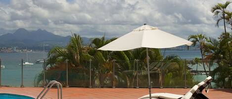 A poolside area with lounge chairs and an umbrella, overlooking a body of water and mountains.