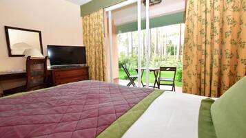 Habitación de hotel con cama, televisión, escritorio con lámpara y vista a un jardín a través de una puerta corrediza de vidrio.