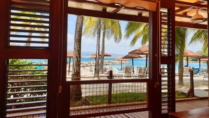 A wooden-framed window with horizontal blinds, overlooking a beach with palm trees and umbrellas. - Hotel Bakoua Les Trois Ilets (Trois-Ilets)