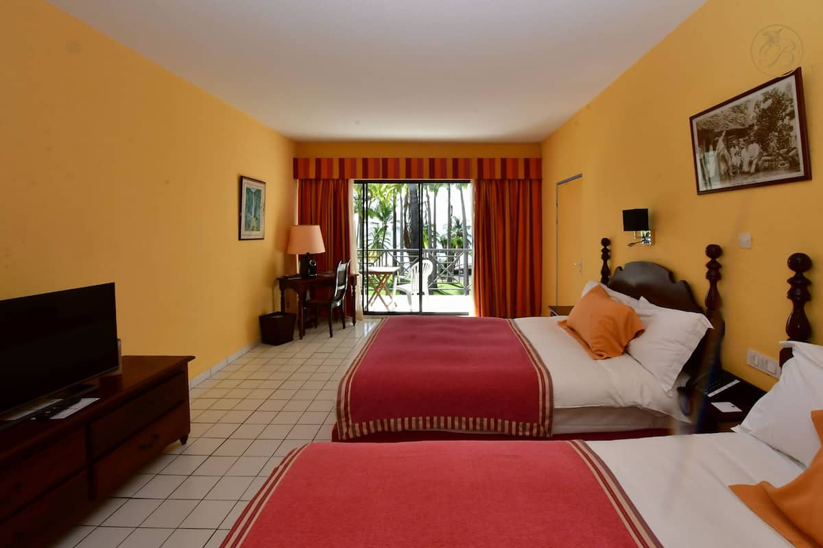 Habitación de hotel con dos camas, televisión, escritorio con lámpara y balcón con vistas a palmeras.