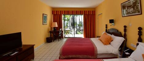 A hotel room with two beds, a television, a desk with a lamp, and a balcony with a view of palm trees.