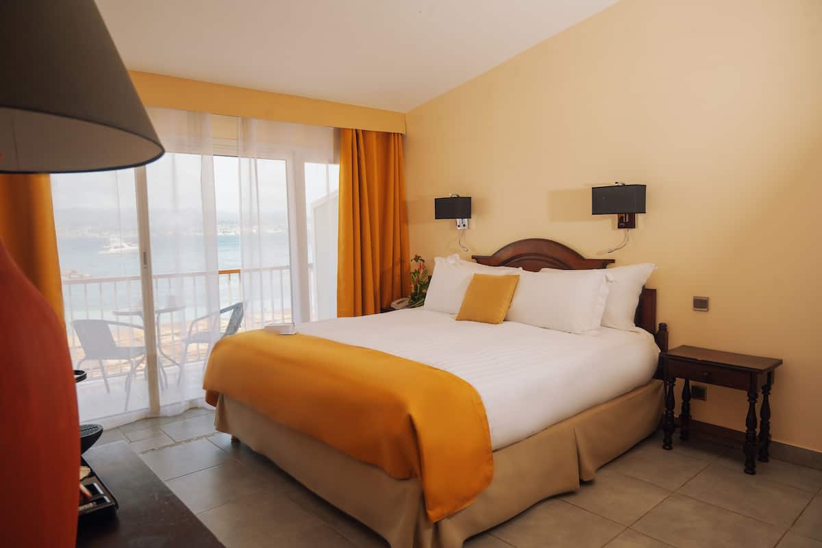 Habitación de hotel con una cama grande, una mesita de noche, una lámpara, una ventana con cortinas y vistas al mar.