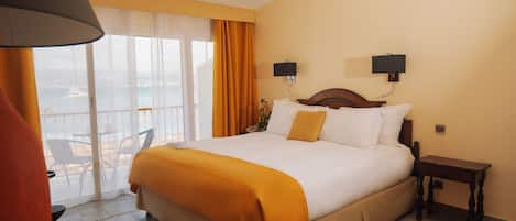 Habitación de hotel con una cama grande, una mesita de noche, una lámpara, una ventana con cortinas y vistas al mar.