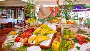A buffet table with various fruits and vegetables, a straw hat, and tropical plants. - Hotel Bakoua Les Trois Ilets (Trois-Ilets)