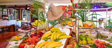 A buffet table with various fruits and vegetables, a straw hat, and tropical plants.