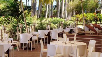 An outdoor dining area with round tables and white chairs, surrounded by lush greenery and palm trees.