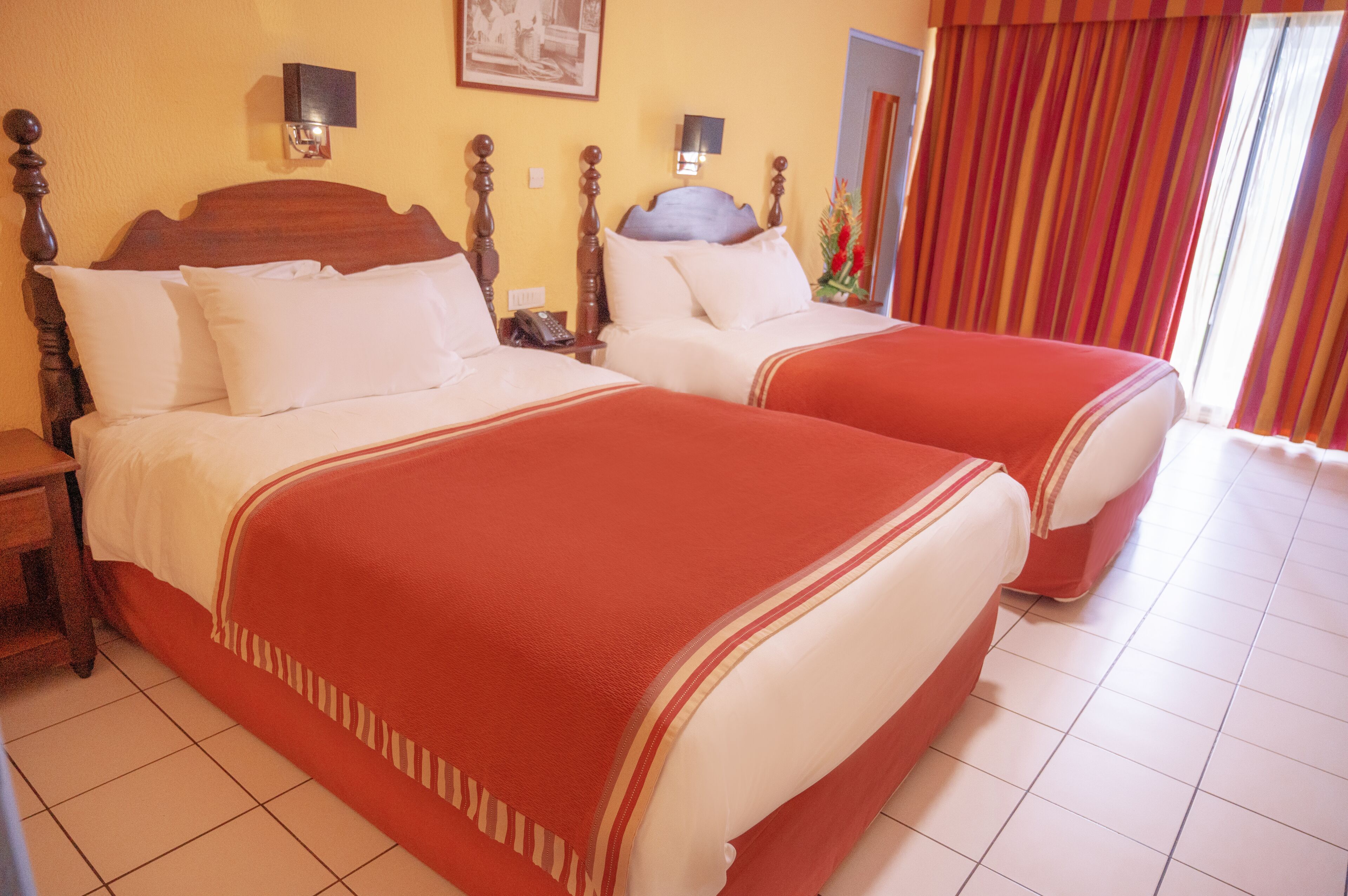 A hotel room with two beds, each with a wooden headboard, white bedding, and red accents. There are bedside tables with lamps, a wall-mounted telephone, and a framed picture above the beds.
