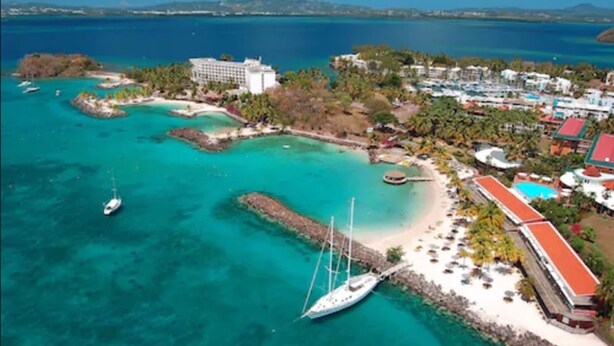 An aerial view of a coastal resort with a large hotel, clear turquoise waters, and a sandy beach. - Hotel Bakoua Les Trois Ilets (Trois-Ilets)