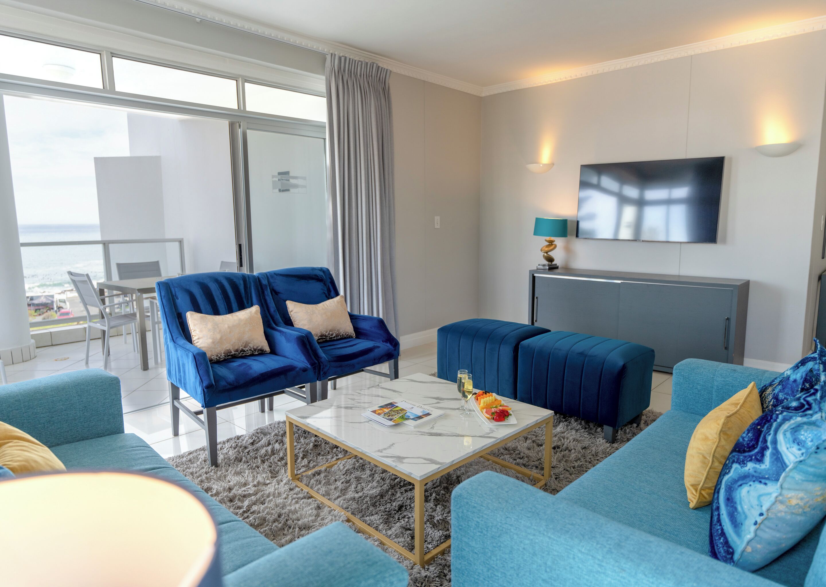A modern living room with a blue sofa, a coffee table, and a TV. There is a balcony with a view of the ocean.