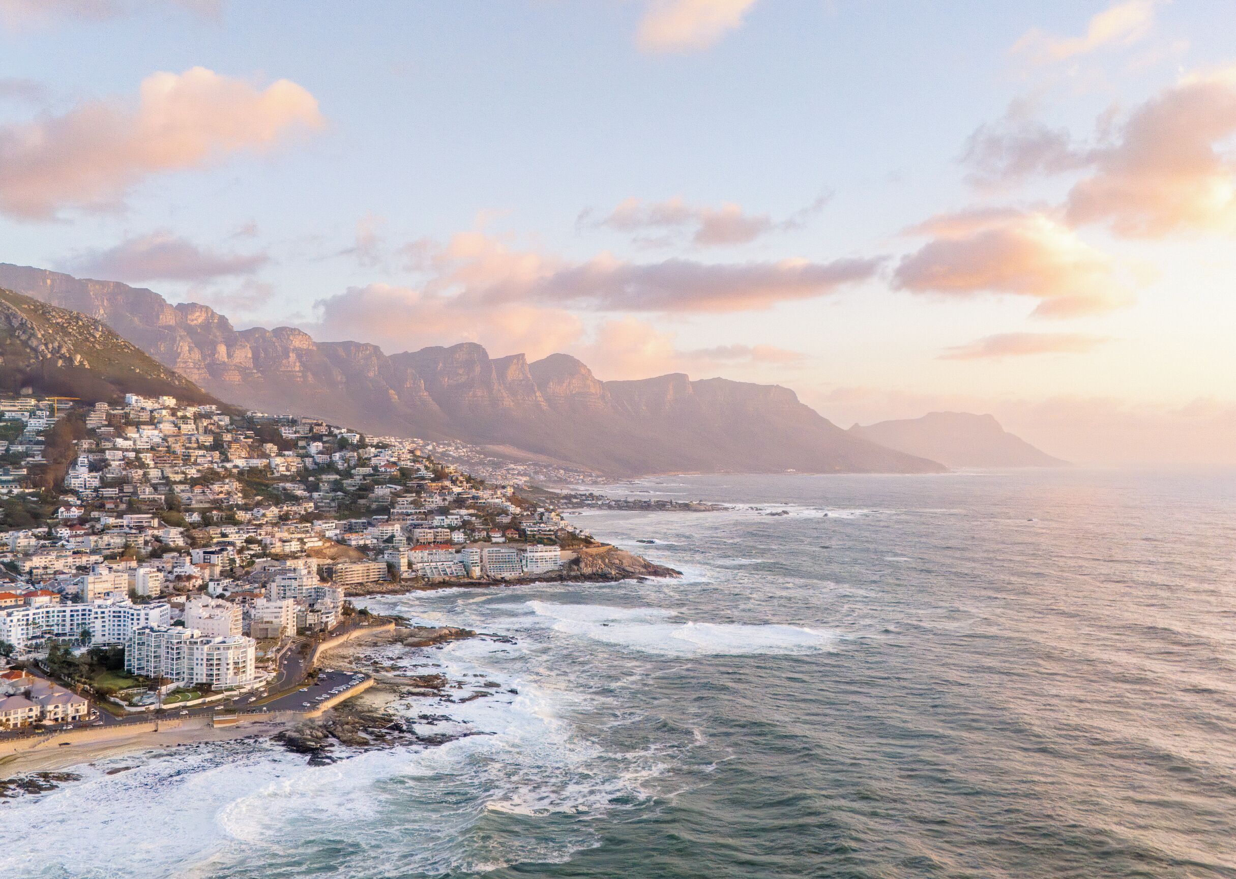 A coastal cityscape with buildings along the shoreline, a rocky coastline, and mountains in the background.