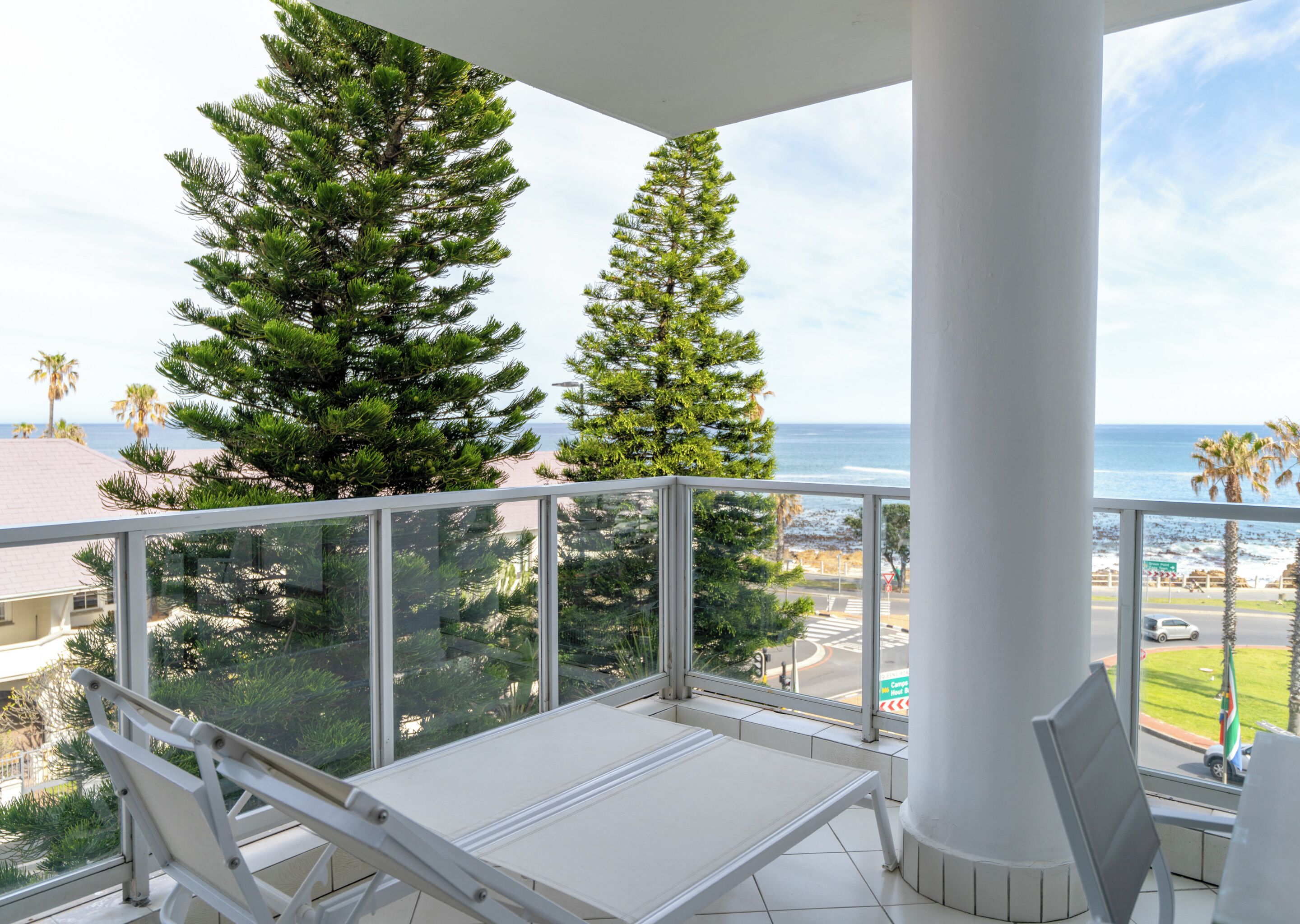A balcony with a white table and chairs, overlooking a beach and ocean with tall pine trees.