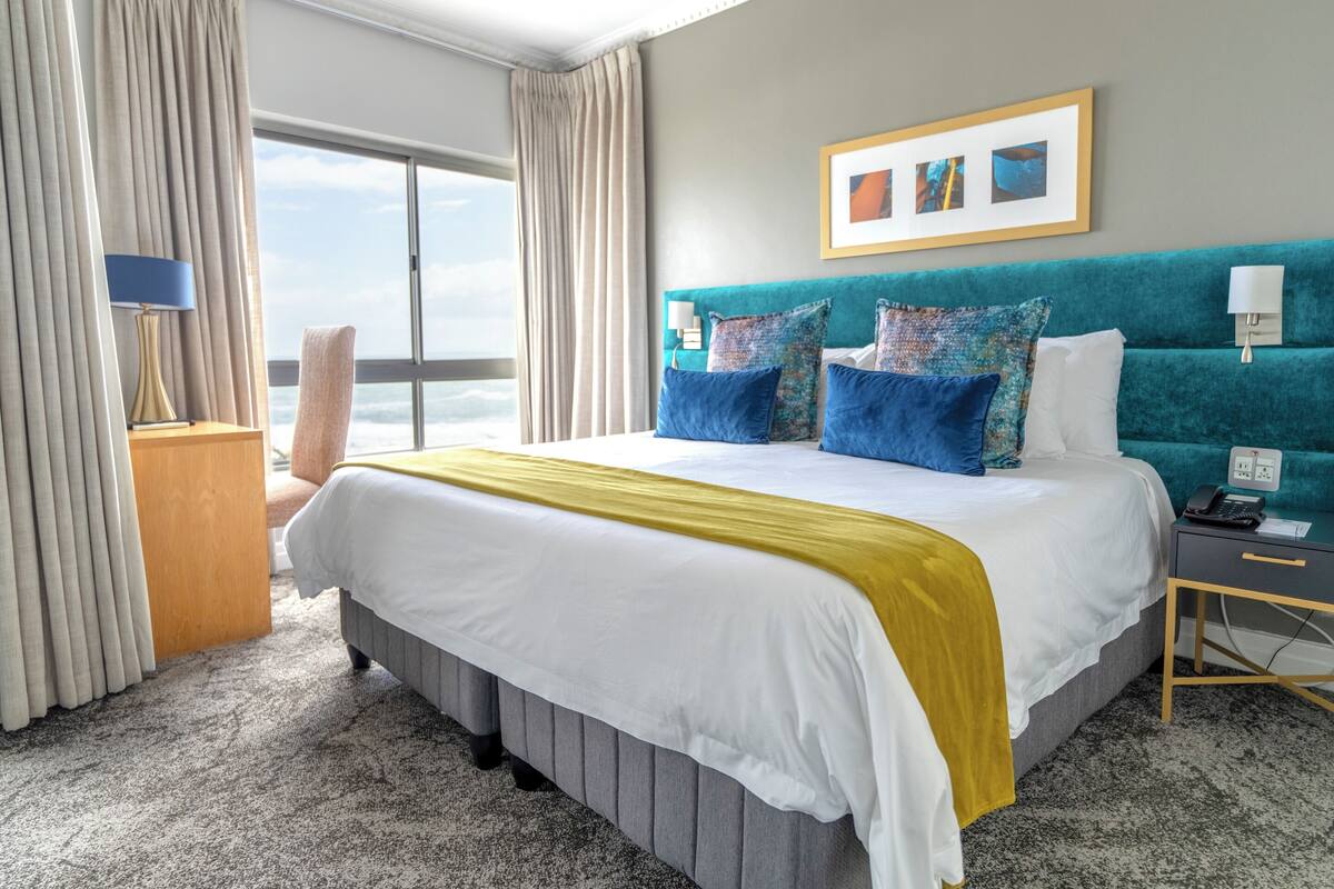 A hotel room with a large bed, a view of the ocean, a teal headboard, and a framed picture on the wall.