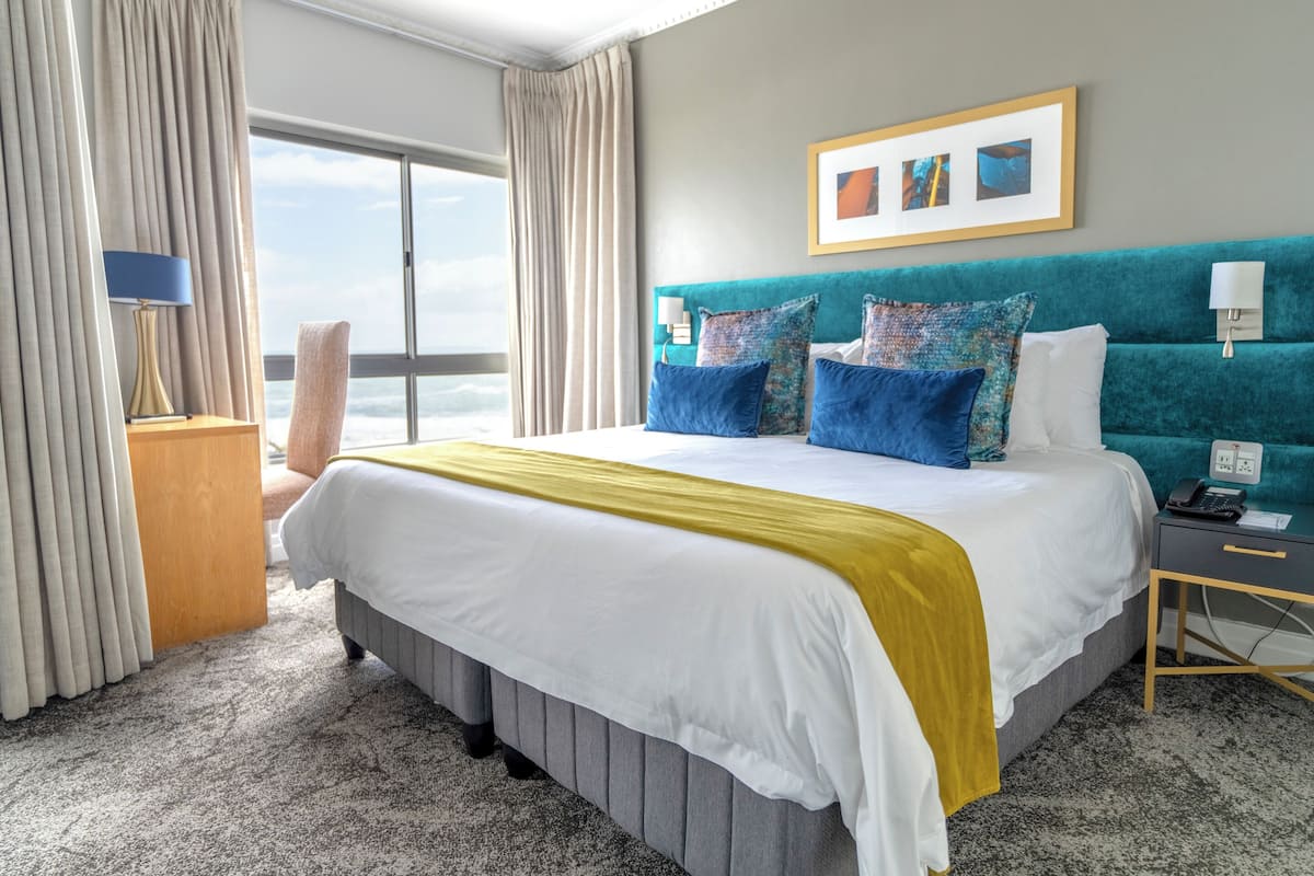 A hotel room with a large bed, a view of the ocean, a teal headboard, and a framed picture on the wall.