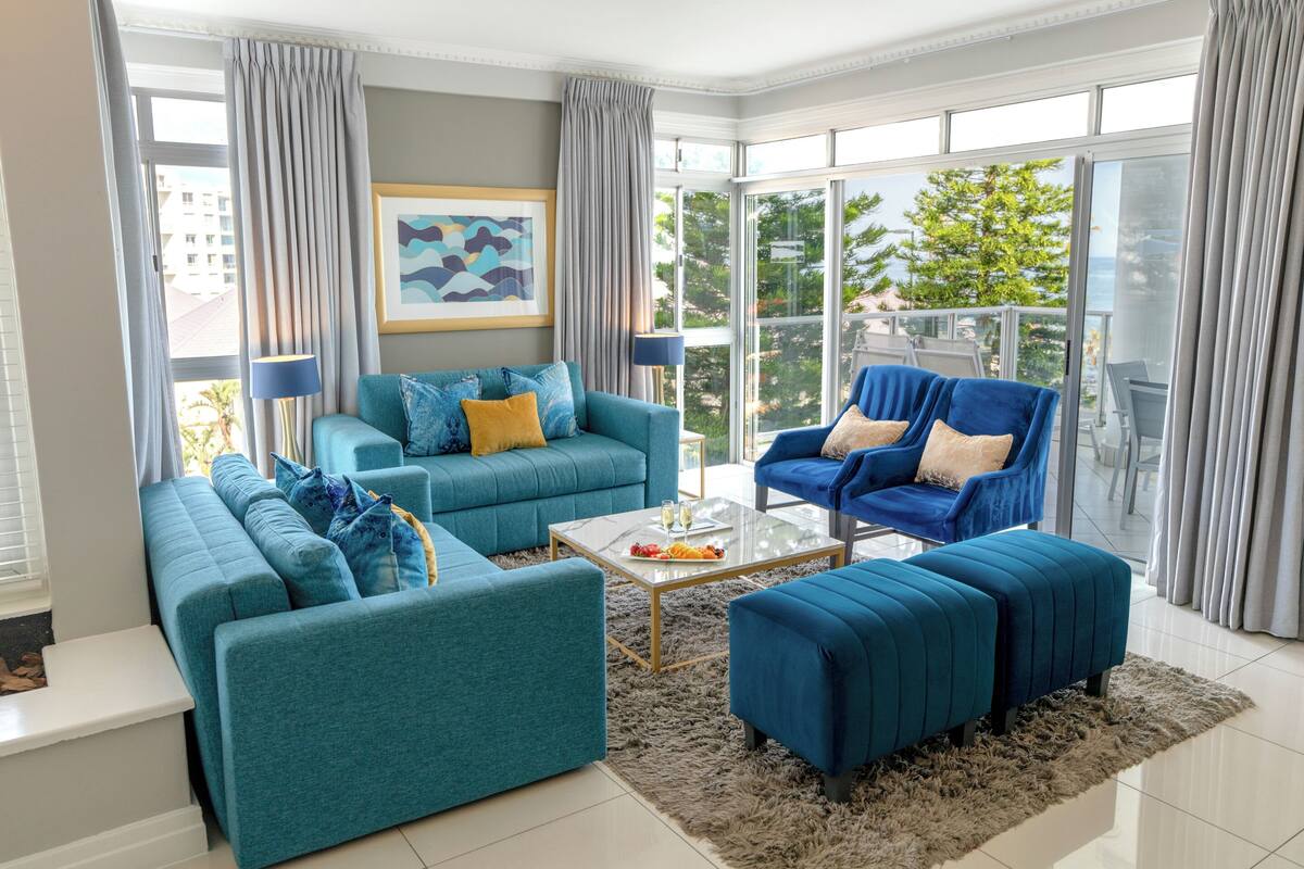 A modern living room with teal sofas, a glass coffee table, and a view of greenery through large windows.