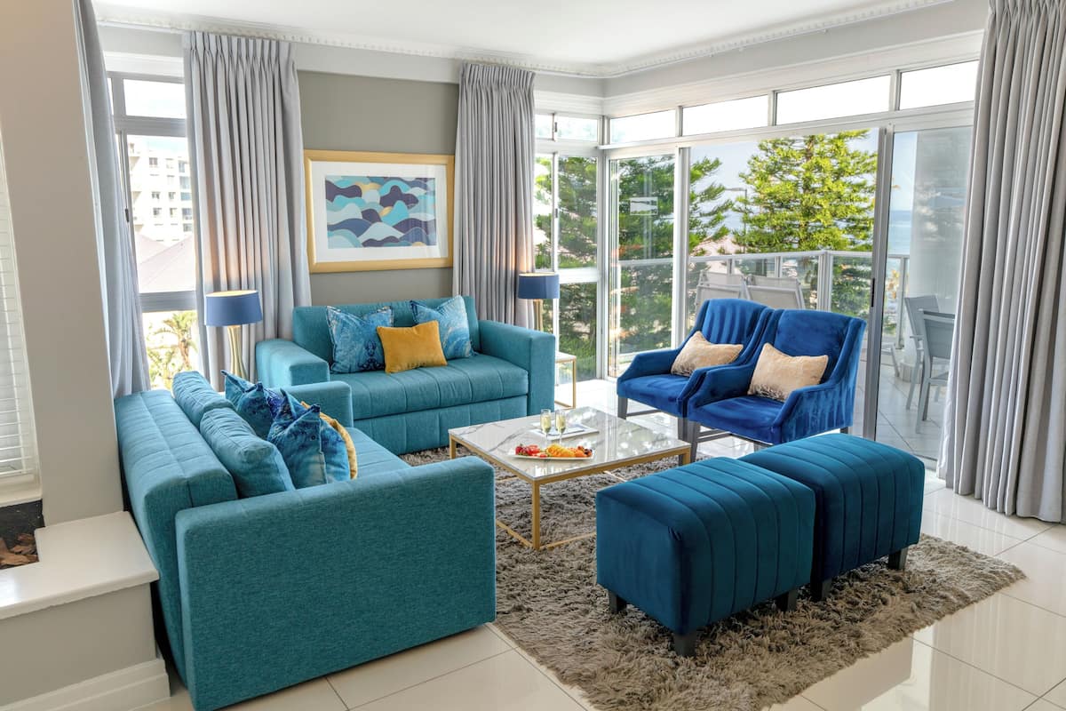 A modern living room with teal sofas, a glass coffee table, and a view of greenery through large windows.