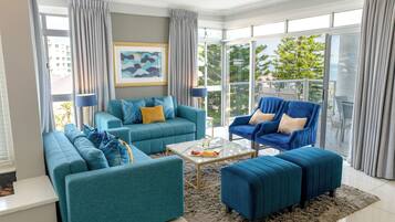 A modern living room with teal sofas, a glass coffee table, and a view of greenery through large windows.