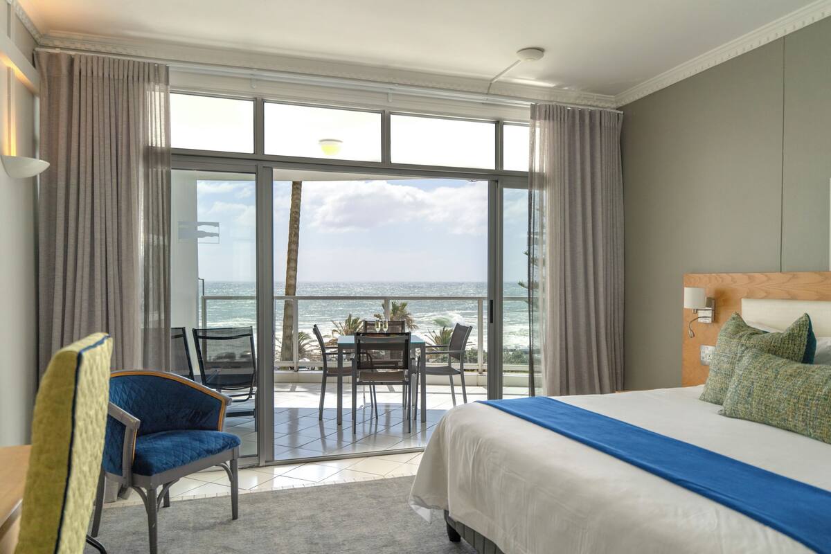 A hotel room with a bed, a chair, a table, and a view of the ocean.