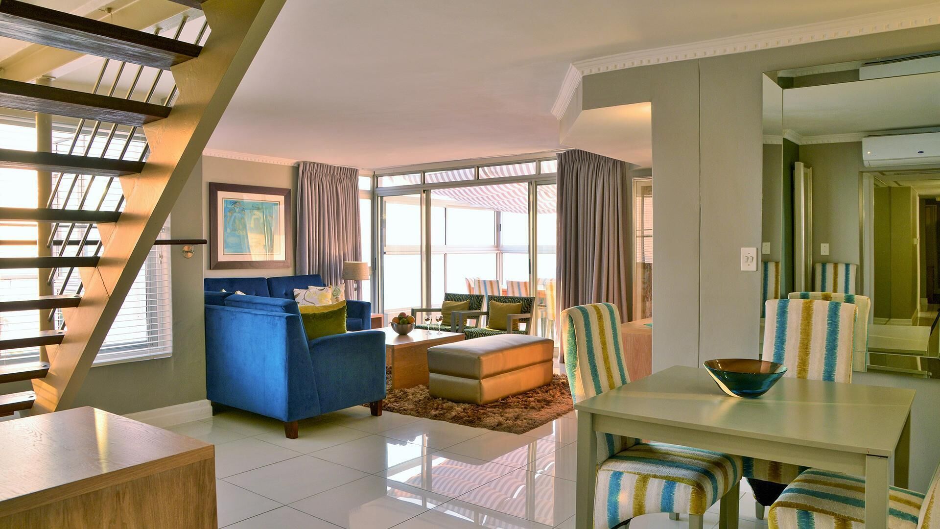 A modern living room with a blue sofa, a dining table with striped chairs, and a staircase.