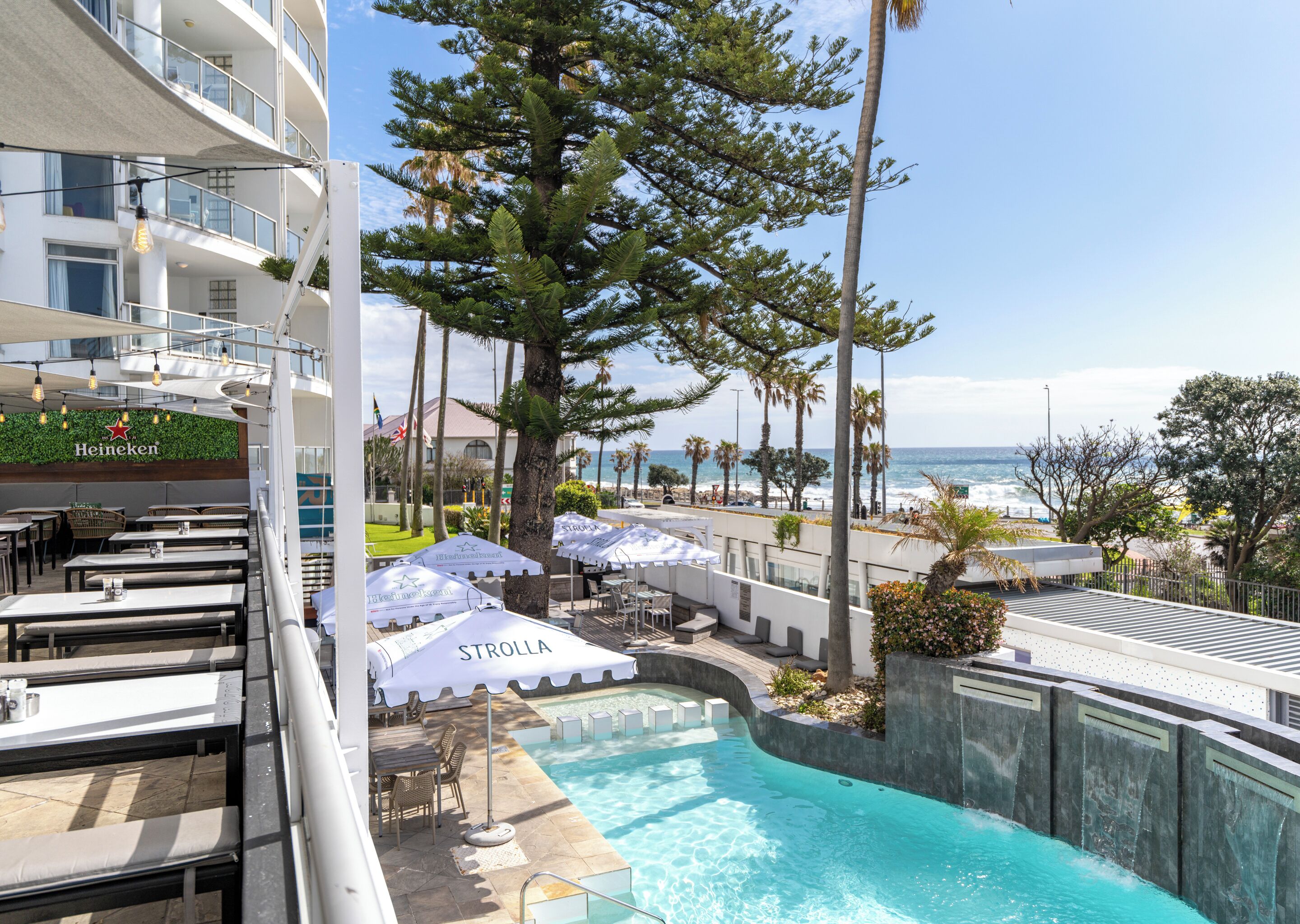 A rooftop pool area with lounge chairs and umbrellas, overlooking a beach and ocean view.
