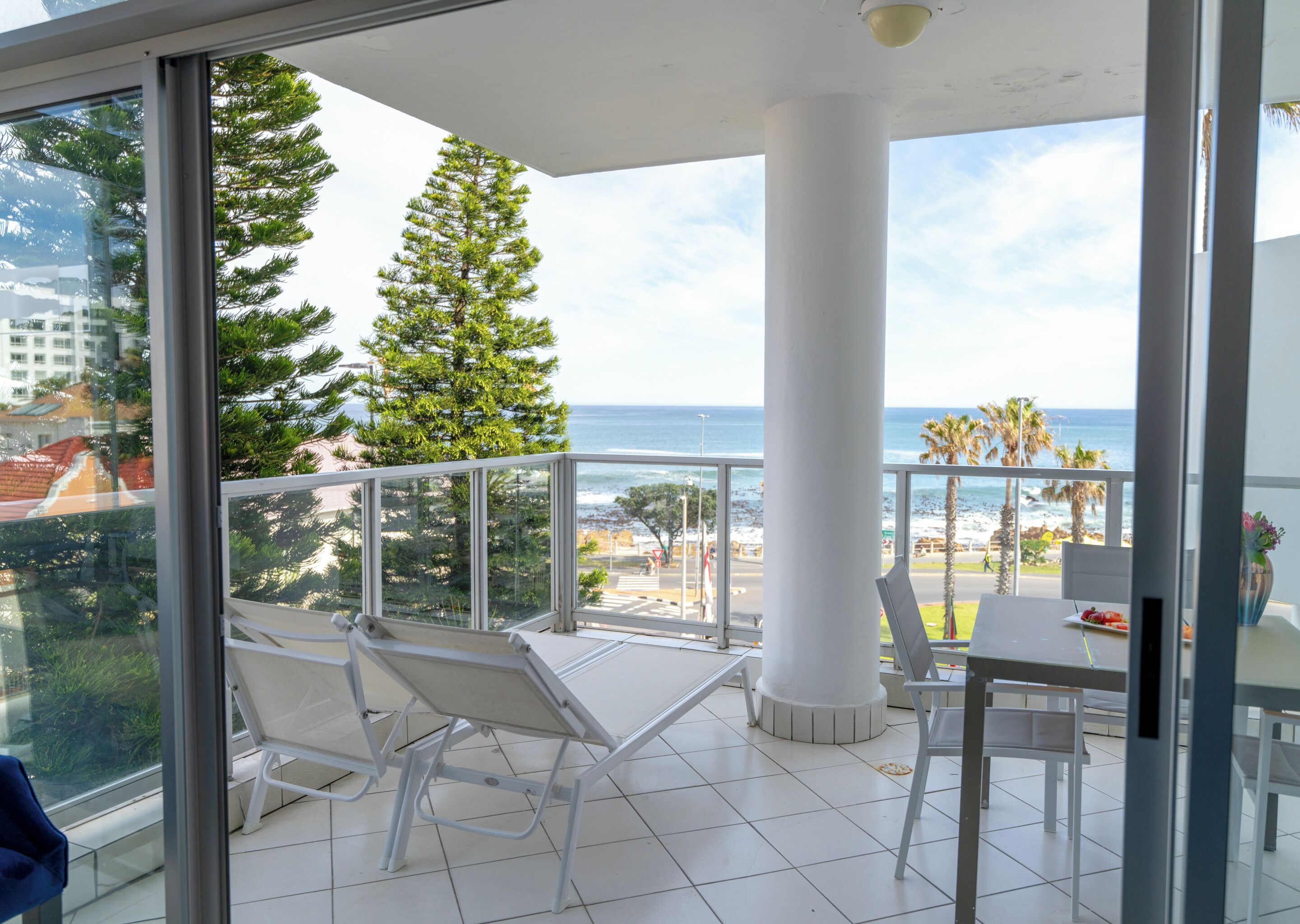 A balcony with a view of the ocean, a white chair, a dining table, and a potted plant.