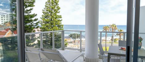 A balcony with a view of the ocean, a white chair, a dining table, and a potted plant.