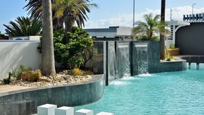 A swimming pool with a waterfall feature, surrounded by palm trees and a modern building. - Peninsula All-Suite Hotel by Dream Resorts (Cape Town)