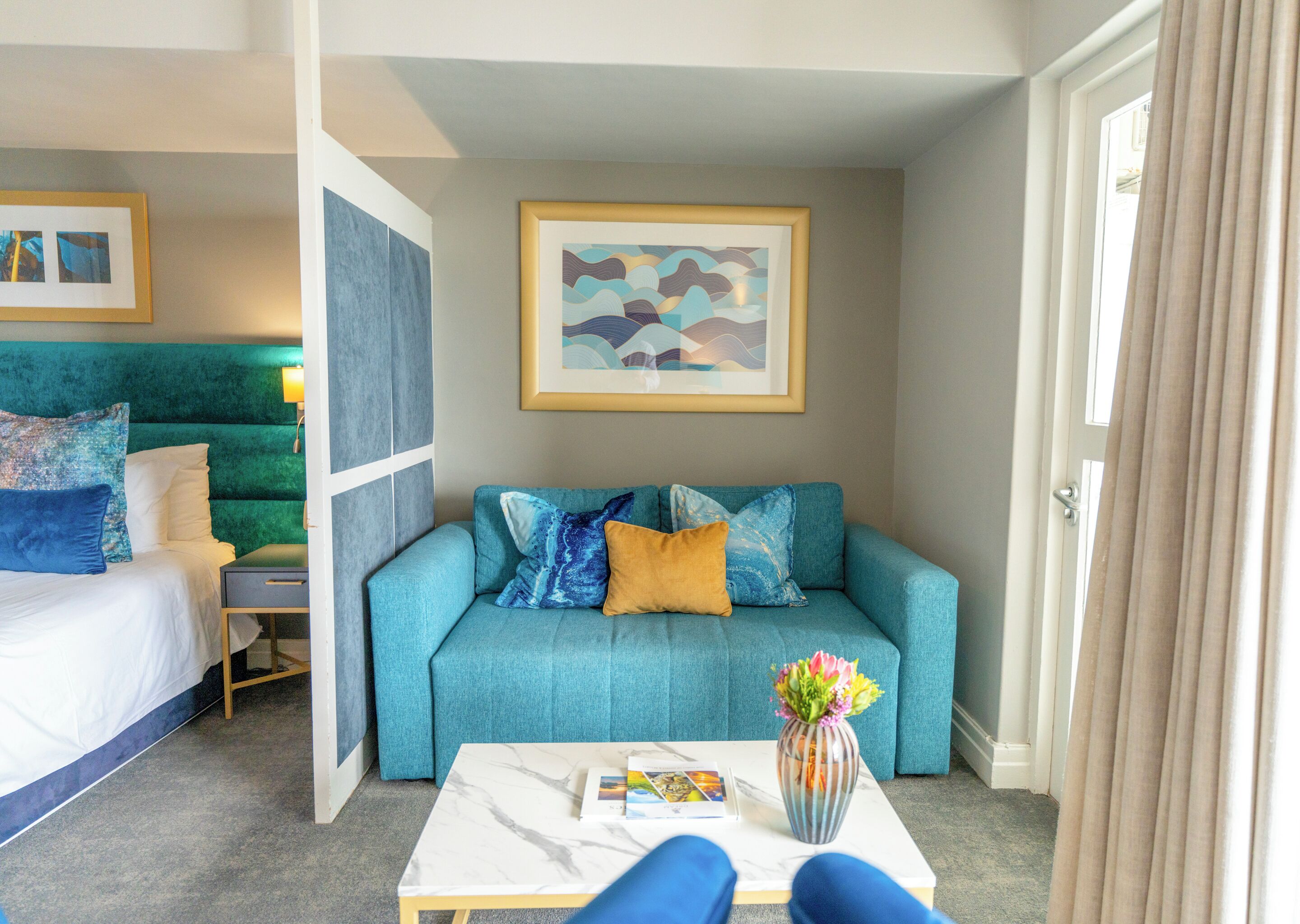 A hotel room with a bed, a blue sofa, a coffee table with a vase, and a framed picture on the wall.