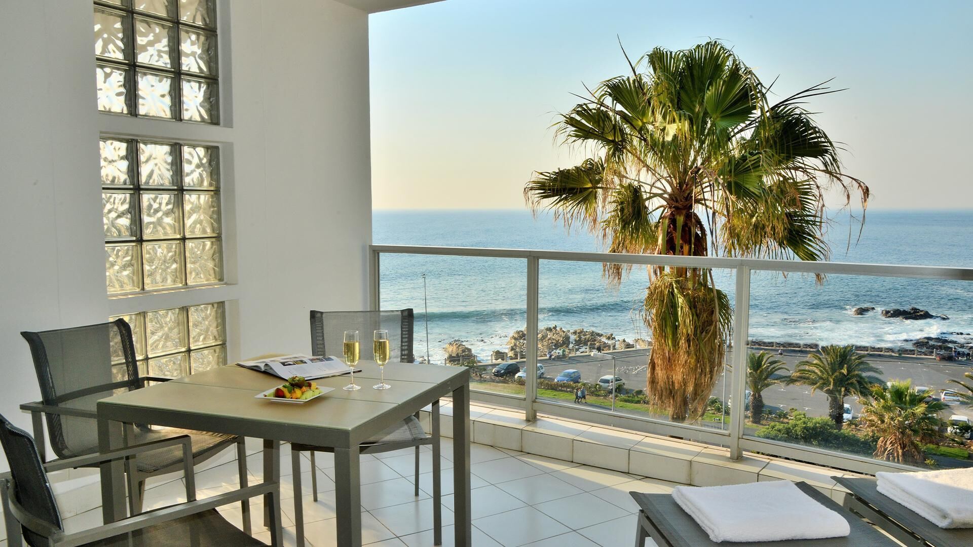 A balcony with a table, chairs, and a view of the ocean and palm trees.