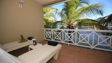 A balcony with two lounge chairs, a small table with a drink, and a view of palm trees and water.