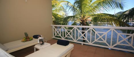 Un balcon avec deux fauteuils, une petite table avec une boisson, et une vue sur des palmiers et l’eau.