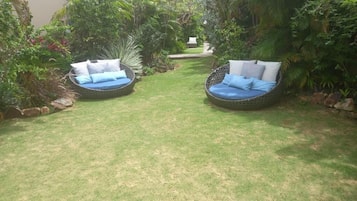 Two wicker outdoor seating areas with cushions, surrounded by lush greenery and a well-maintained lawn.