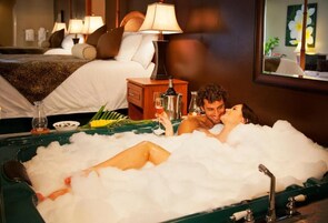 Een koppel ontspant in een bad vol bubbels, met een bed en lampen op de achtergrond.
