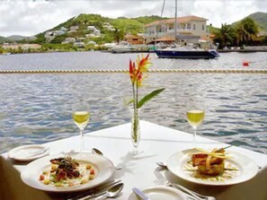 A table set with plates of food and two glasses of white wine, overlooking a body of water with boats and a hillside in the background. - Harmony Marina Suites (Gros Islet)