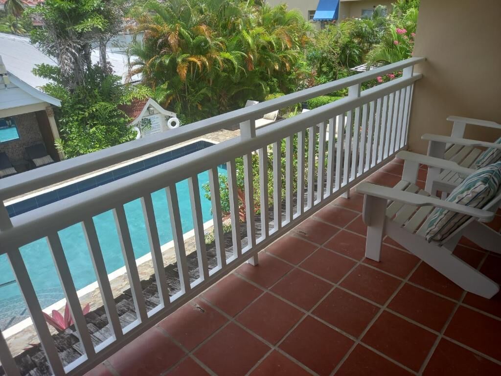 A balcony with a white railing, a pool, and a view of greenery and buildings.
