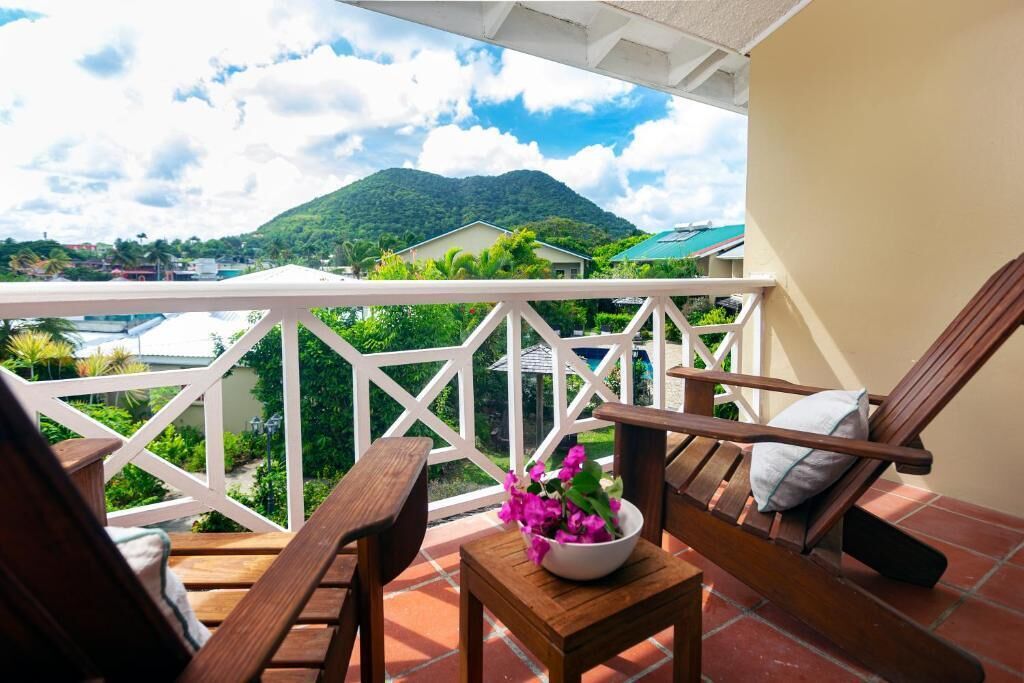 A balcony with wooden chairs and a small table, overlooking a lush green landscape and a mountain.