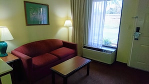 Quarto de hotel com sofá vermelho, uma pequena mesa, luminária, janela com cortinas e radiador.