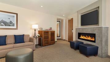 A modern living room with a beige sofa, blue cushions, a round ottoman, a flat-screen TV, a fireplace, and a framed picture on the wall.