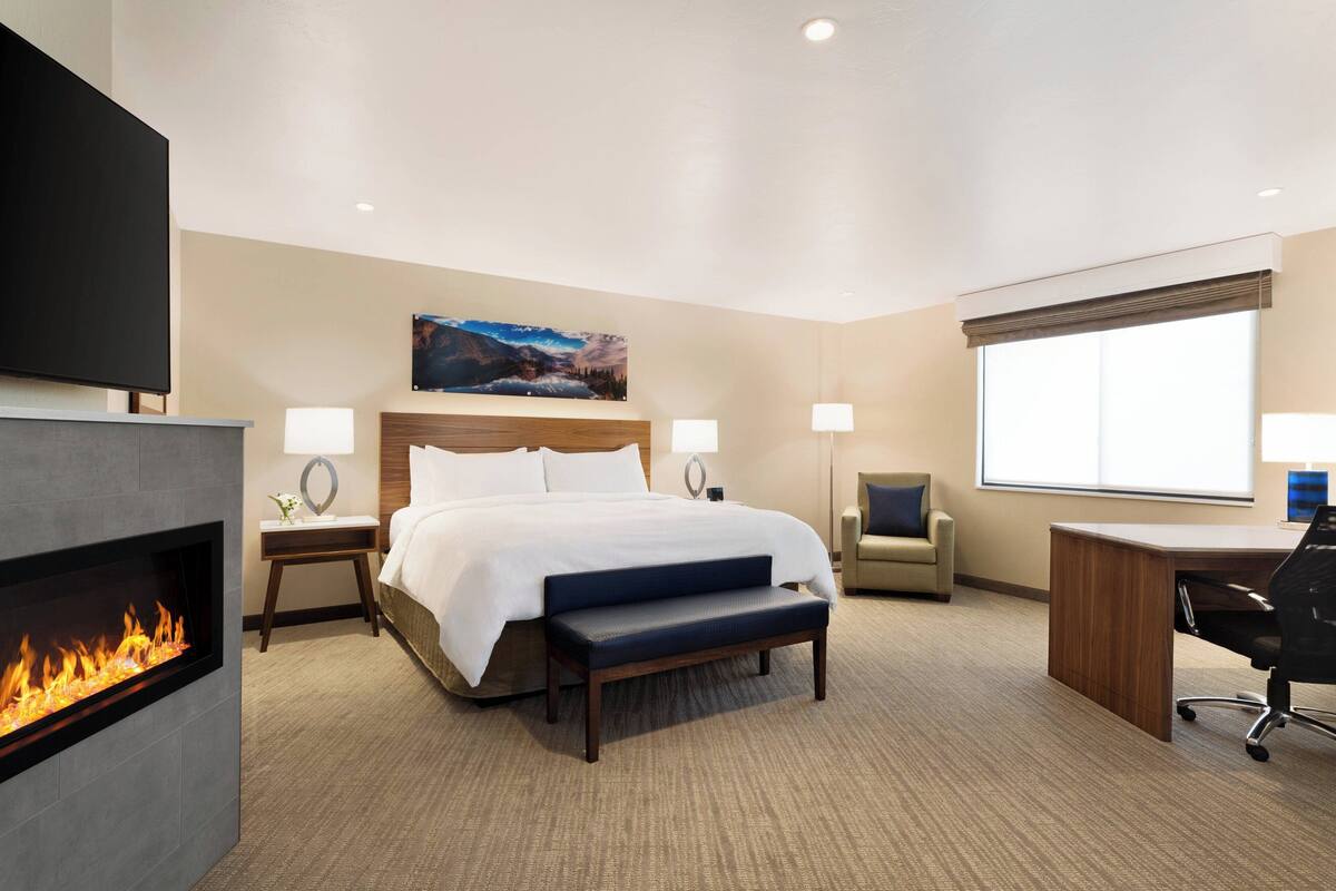 Quarto de hotel com cama, escrivaninha, poltrona, lareira e uma janela grande.