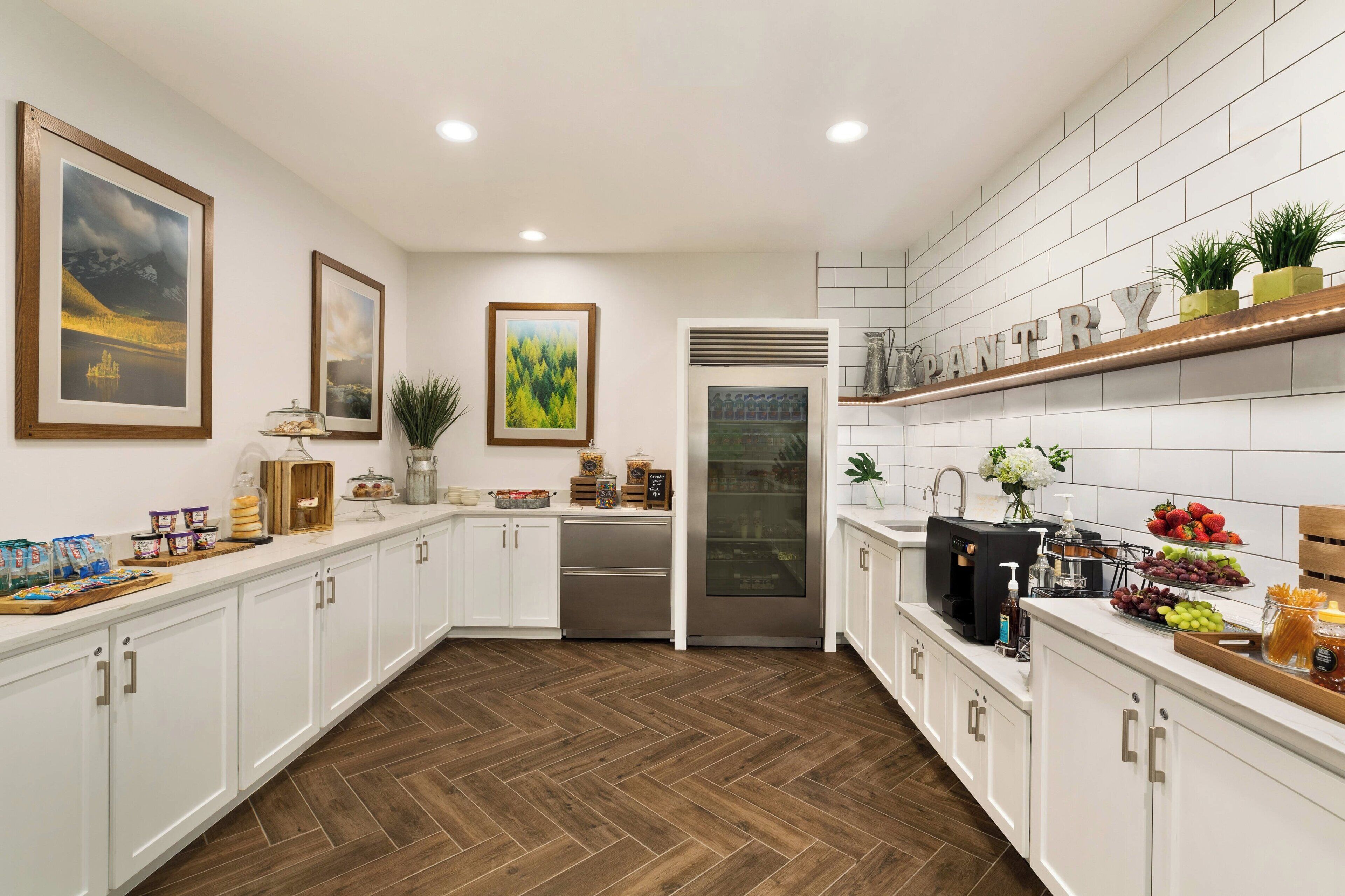 A modern kitchen with white cabinets, a stainless steel refrigerator, and a built-in oven. There are framed pictures on the wall and a shelf with kitchen utensils.