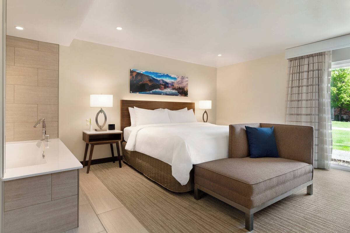 Quarto de hotel com cama, sofá, mesa de cabeceira, abajur e um quadro na parede.