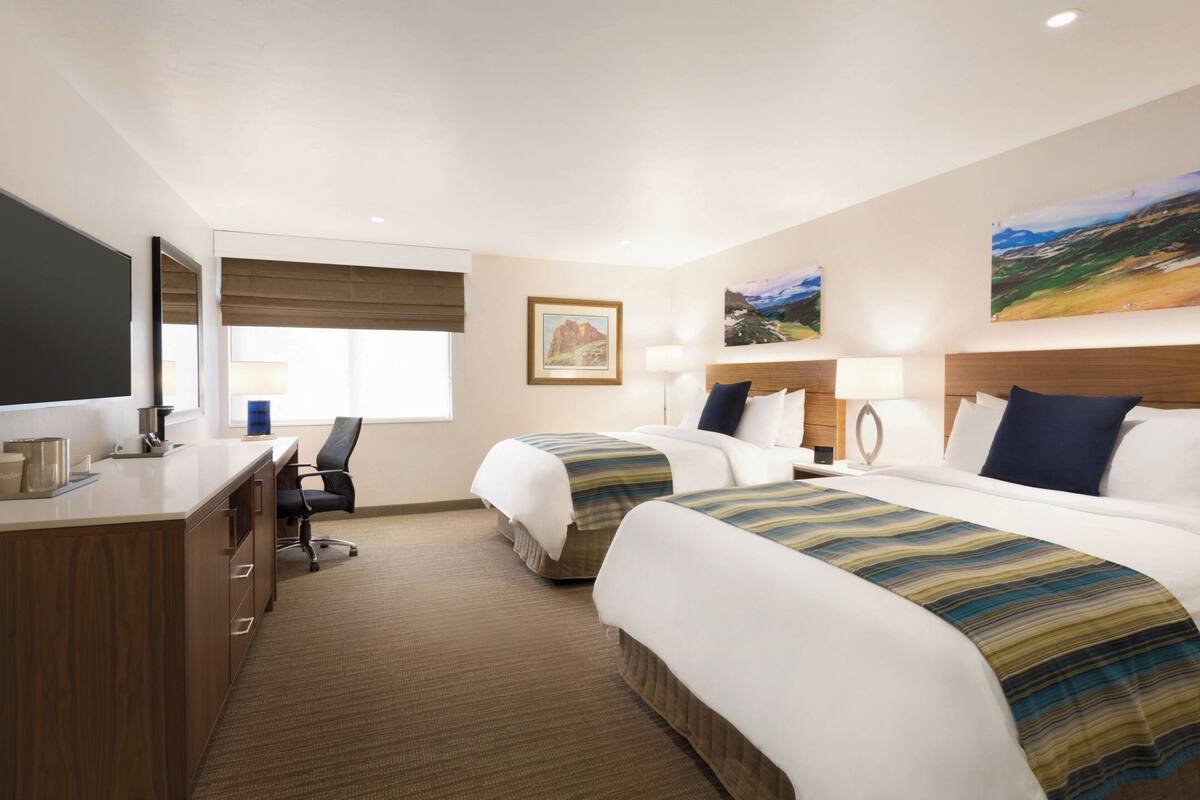 Quarto de hotel com duas camas, uma escrivaninha, uma cadeira e uma janela grande com cortinas.