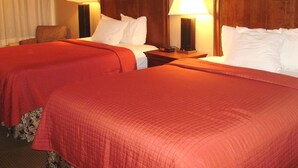 A hotel room with two beds, each with a wooden headboard, white linens, and red bedding. There are two bedside tables with lamps, a chair, and a window with curtains. - Trade Winds Central Inn (Tulsa)