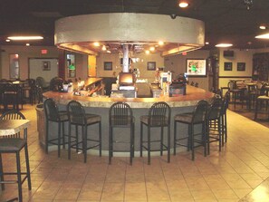 Zona de bar con barra central, varios taburetes y suelo de baldosas.