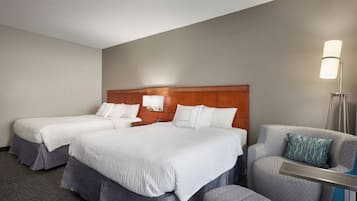 Una habitación de hotel con dos camas, un sillón gris y una mesita de noche con una lámpara.