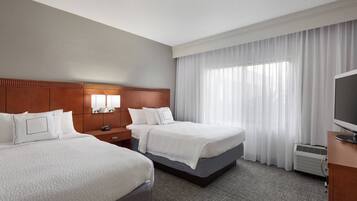 Quarto de hotel com duas camas, cabeceira de madeira, uma janela grande com cortinas finas, televisão de tela plana e aparelho de ar condicionado embutido.