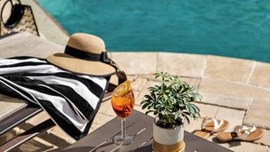 Escena junto a la piscina con té helado en un vaso, un sombrero, una toalla a rayas, una planta en maceta y un par de sandalias.