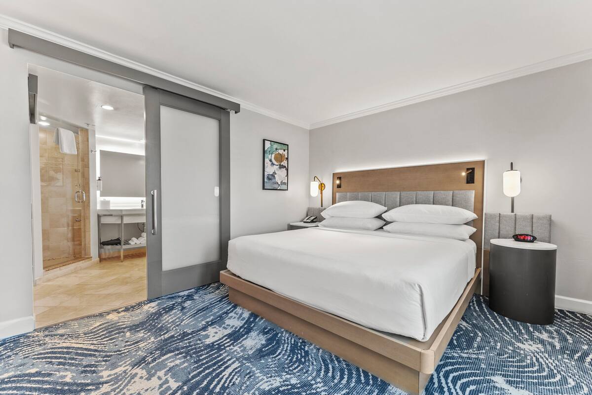 Une chambre d’hôtel moderne dotée d’un grand lit, d’un dressing, d’une salle de bain avec douche et d’une table de chevet avec une lampe.