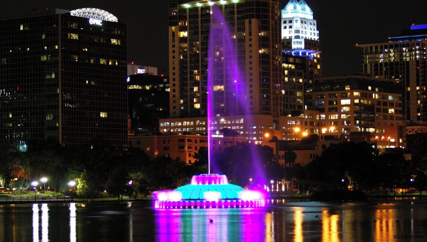 A nighttime cityscape with illuminated buildings and a fountain.