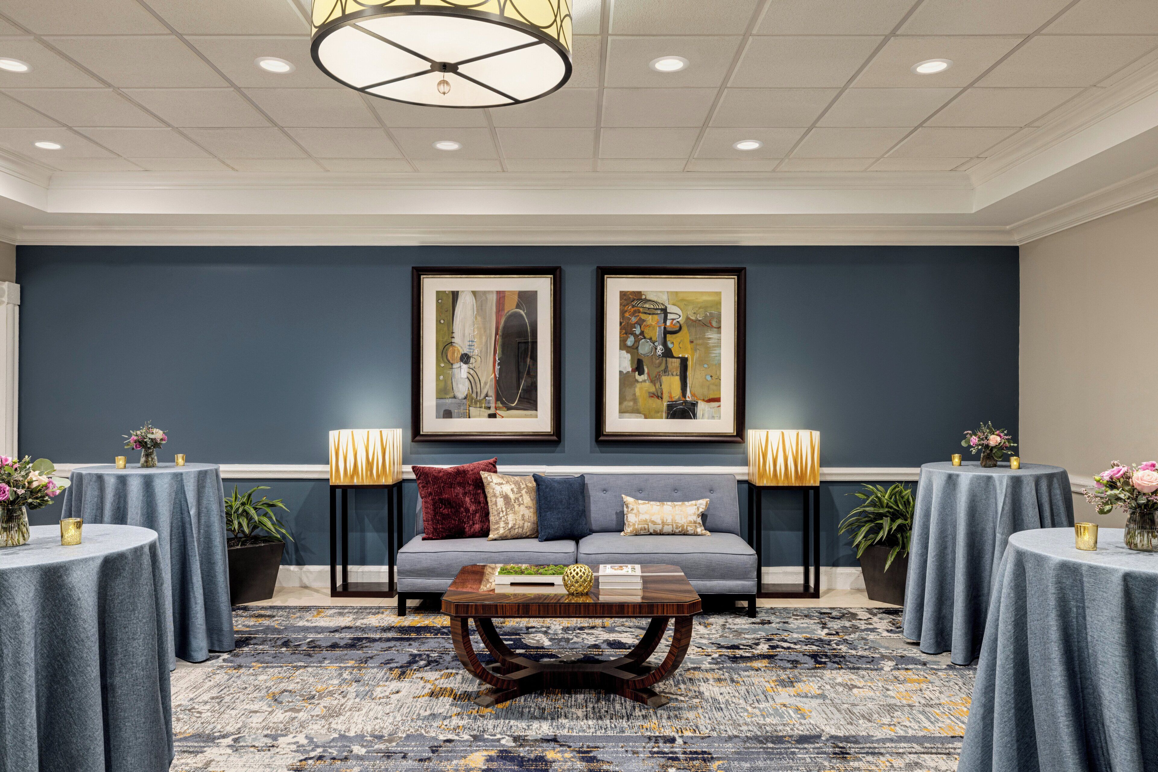 A modern lobby with a grey sofa, two framed artworks, round tables with lamps, and a patterned carpet.