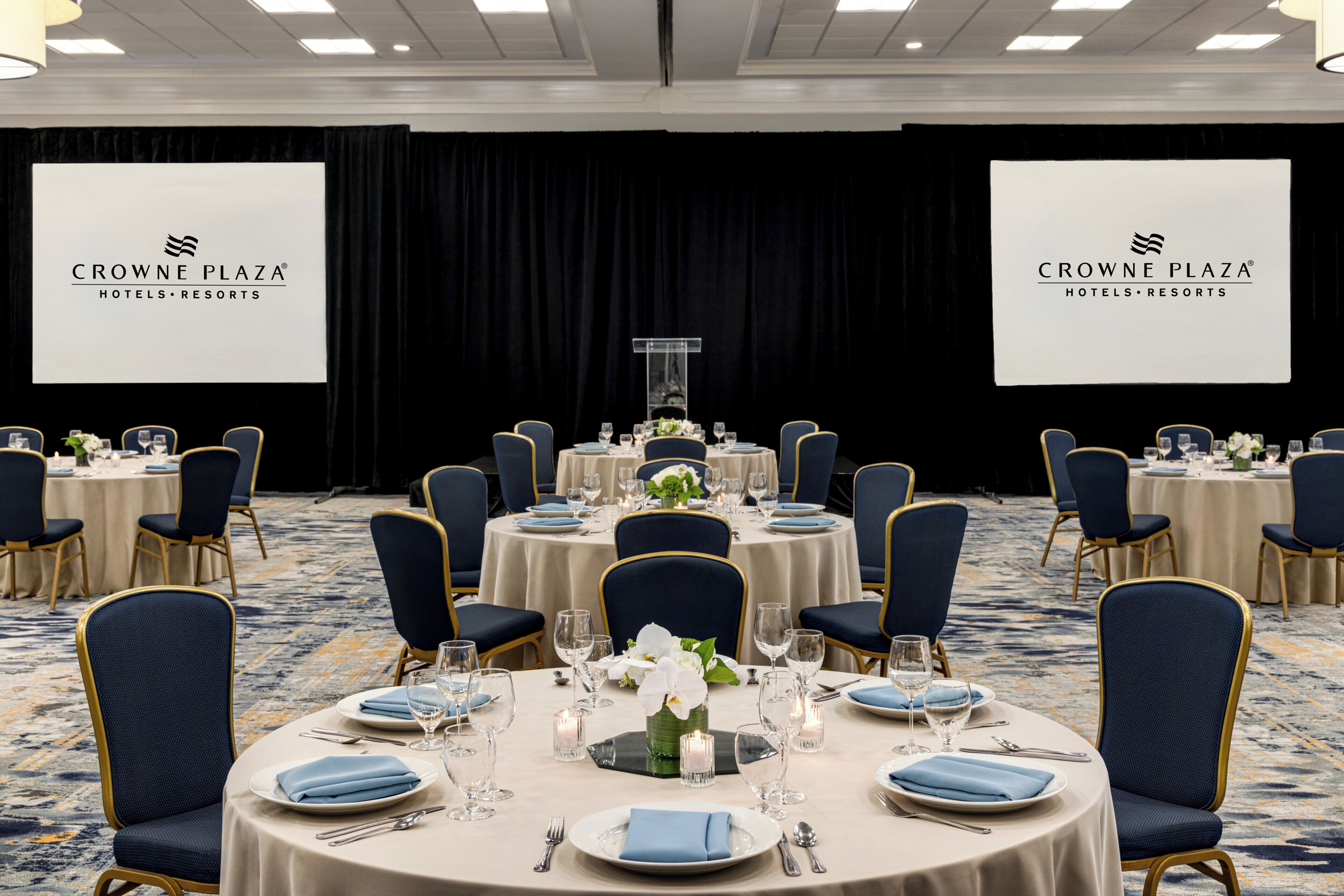 Un salón de conferencias en Crowne Plaza Hotels & Resorts con mesas redondas, sillas azules y piso alfombrado.